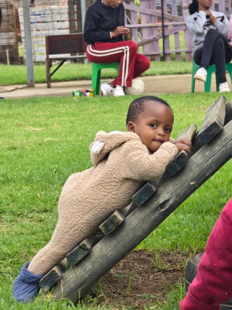 Young child climbing on a wooden log structure at a park while adults supervise in the background on grass
