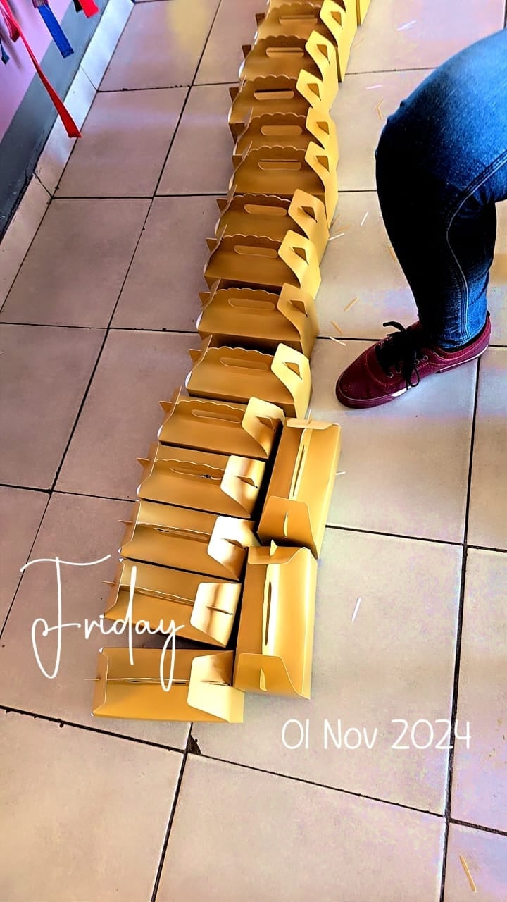 Persons feet in burgundy sneakers standing next to a long line of golden gift boxes arranged on a tiled floor with Friday, 01 Nov 2024 text overlay