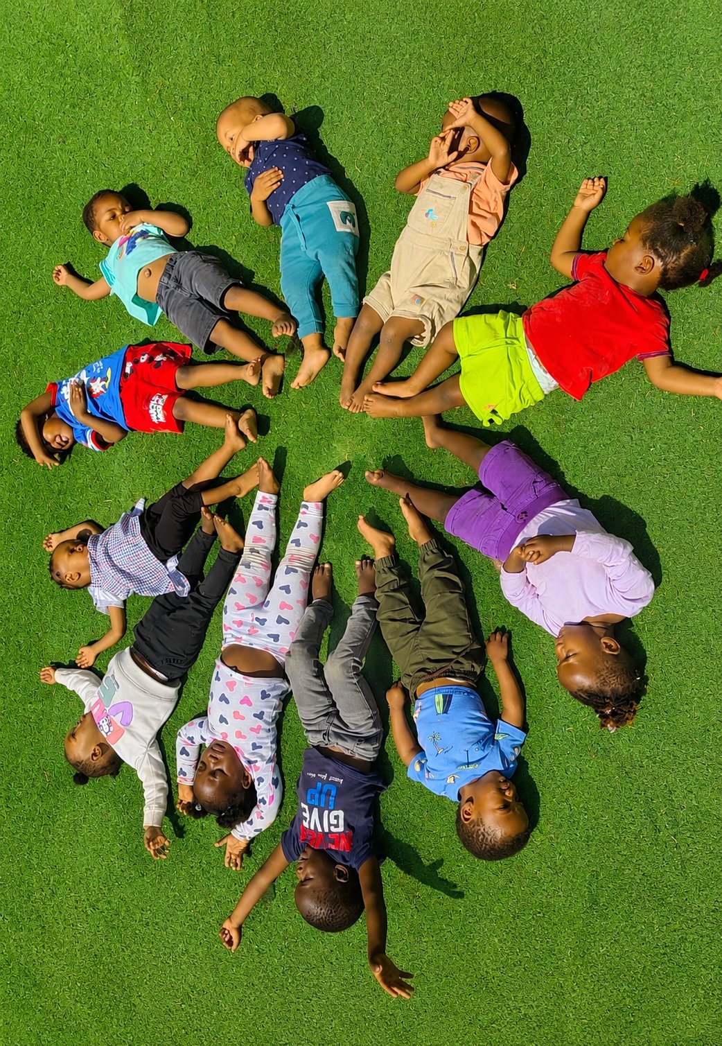 Group of children lying in a circle on green grass, viewed from above, wearing colorful clothing