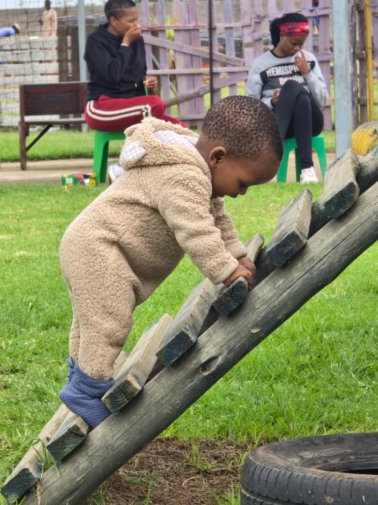 Young child climbing on a wooden log obstacle while other children play in a grassy outdoor recreational area