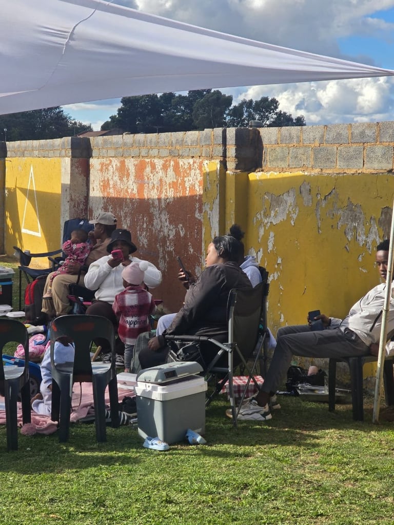 Group of people gathered under a tent canopy in front of weathered yellow and rust-colored metal walls on grass, with coolers and chairs visible