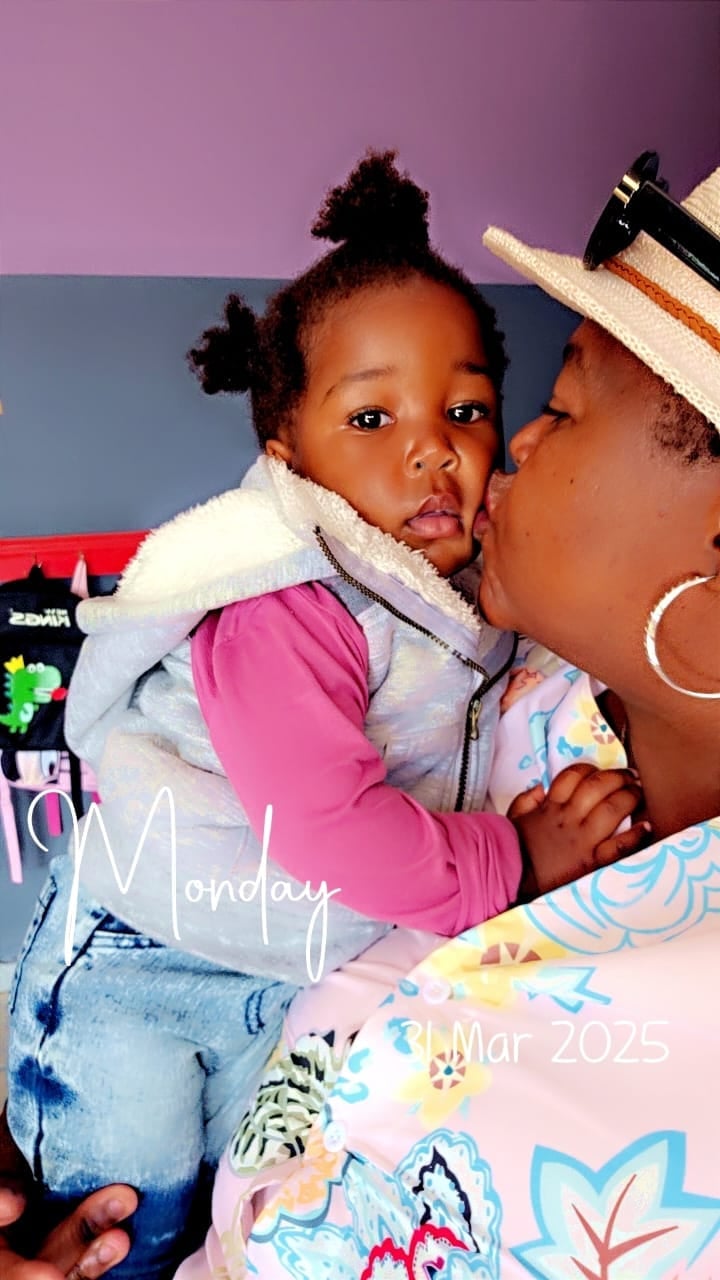 An adult wearing a straw hat kisses a toddler on the cheek. The child wears a pink and gray outfit with distinctive black twisted hair. Text overlay reads Monday and Mar 2025.