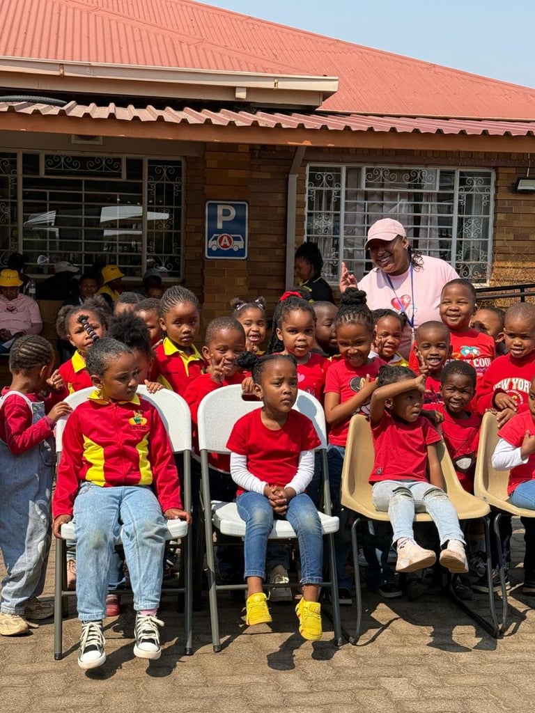 Group of children in red and yellow uniforms seated outside a building with an adult supervisor standing behind them