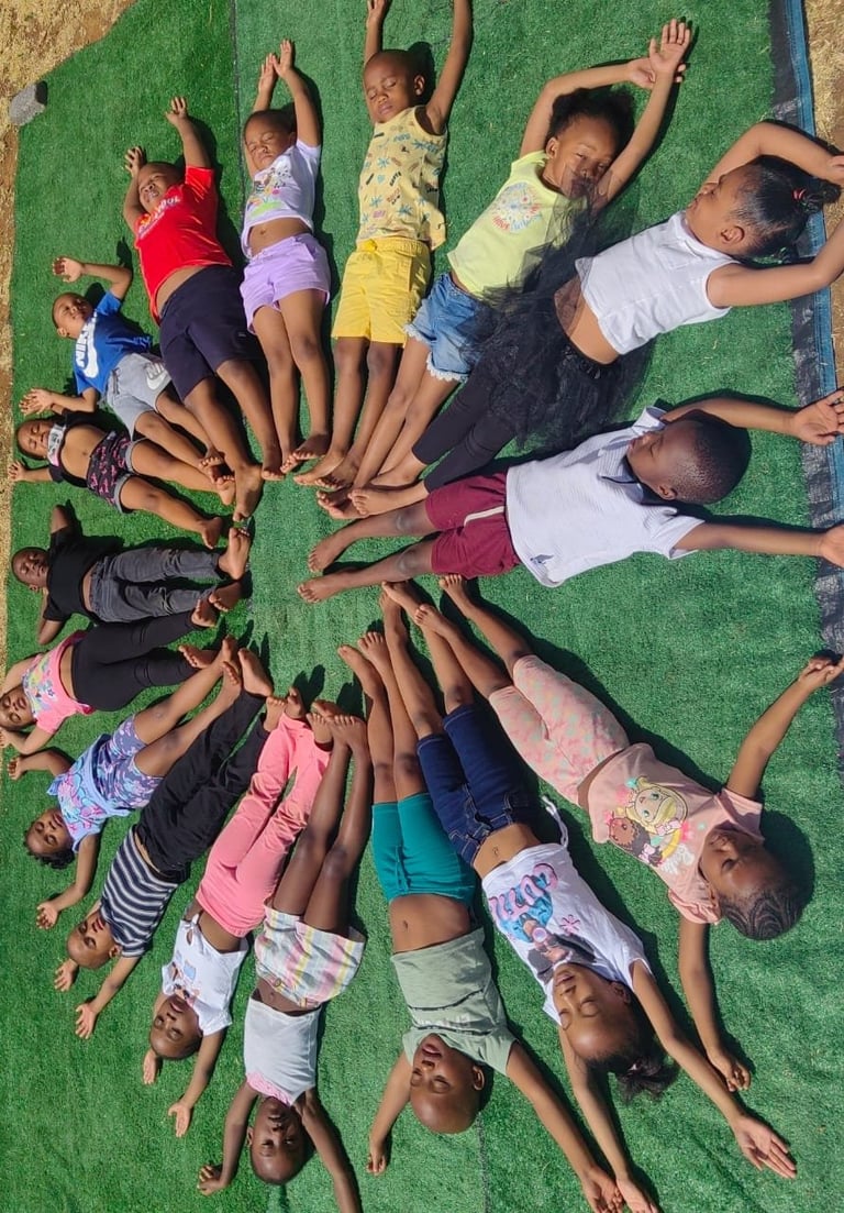 Group of diverse children lying in a circle on green grass, heads toward center, arms raised, smiling at camera