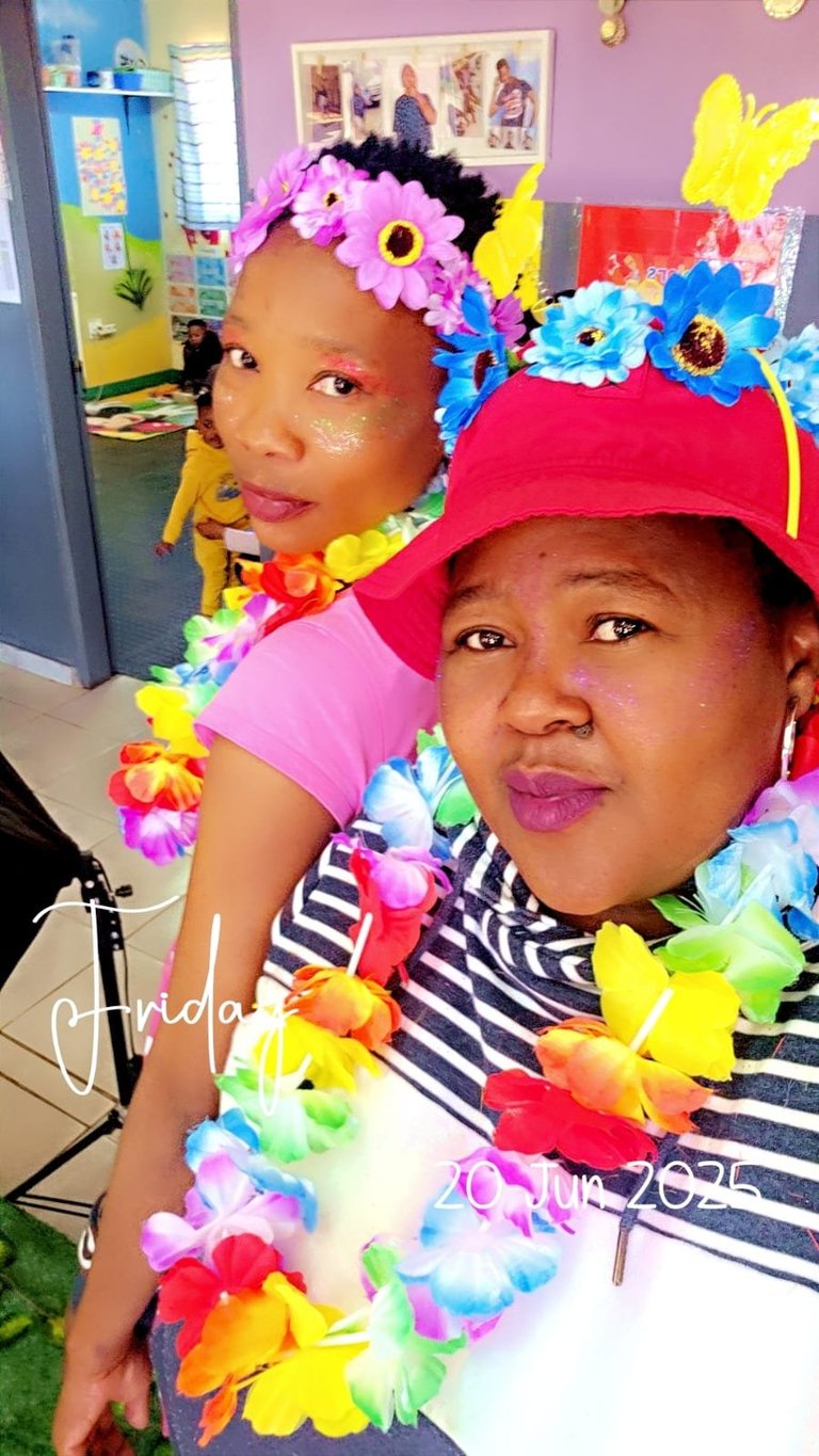 Two children wearing colorful flower crowns and leis, smiling at the camera in a classroom setting with pink walls