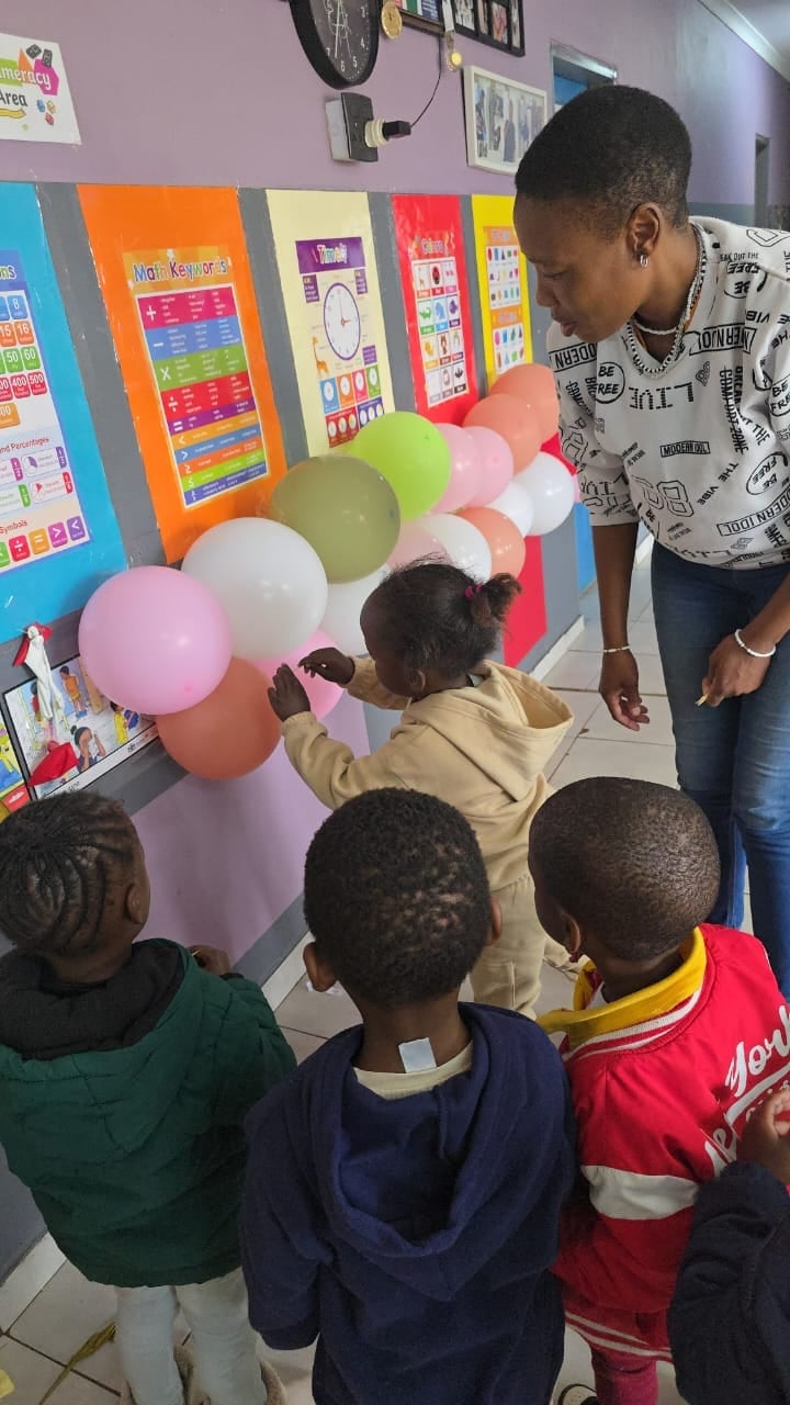 Teacher and elementary students gathered around colorful balloons in a classroom decorated with educational posters and learning charts