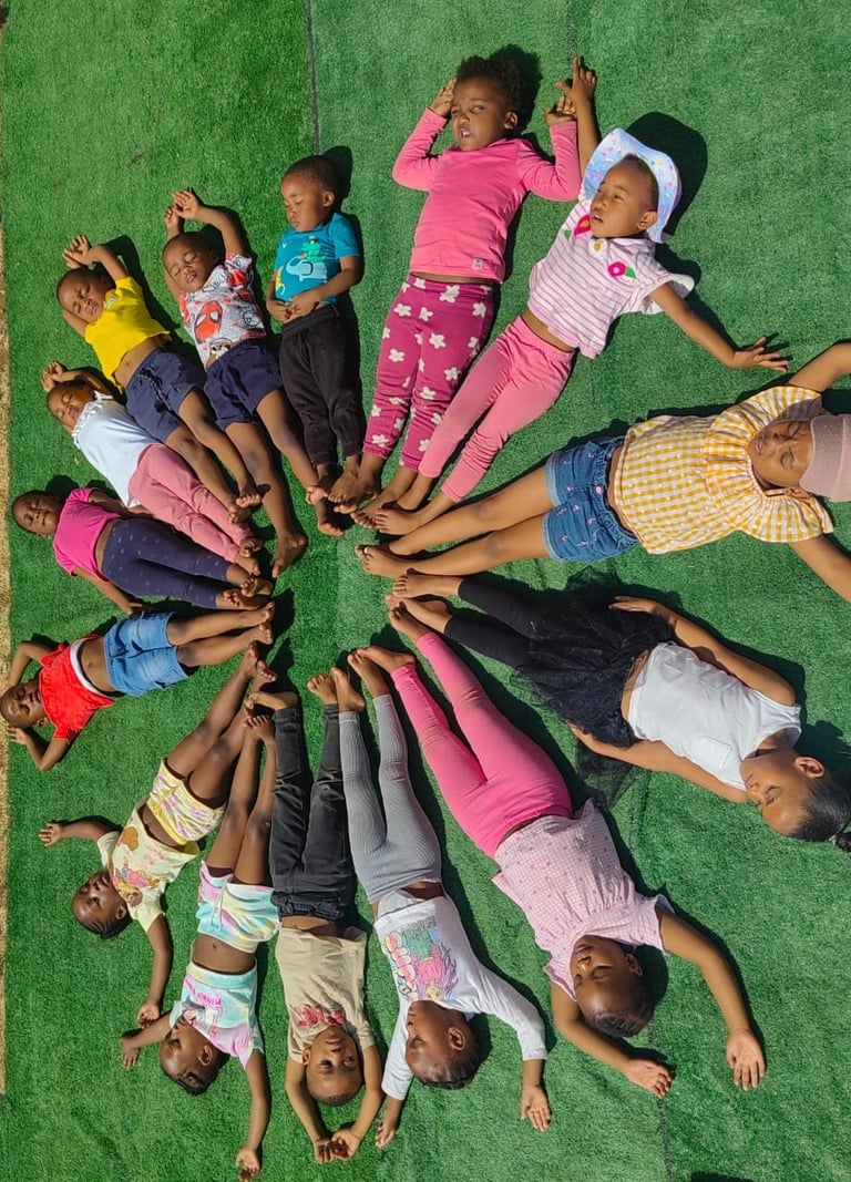 Group of children lying in a circle on green grass with their heads together, looking up at the camera