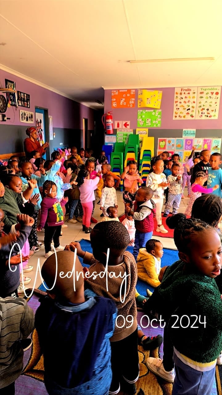 Large group of young children gathered in a colorful classroom with vibrant posters and educational materials on purple and gray walls, dated Wednesday, 09 Oct 2024