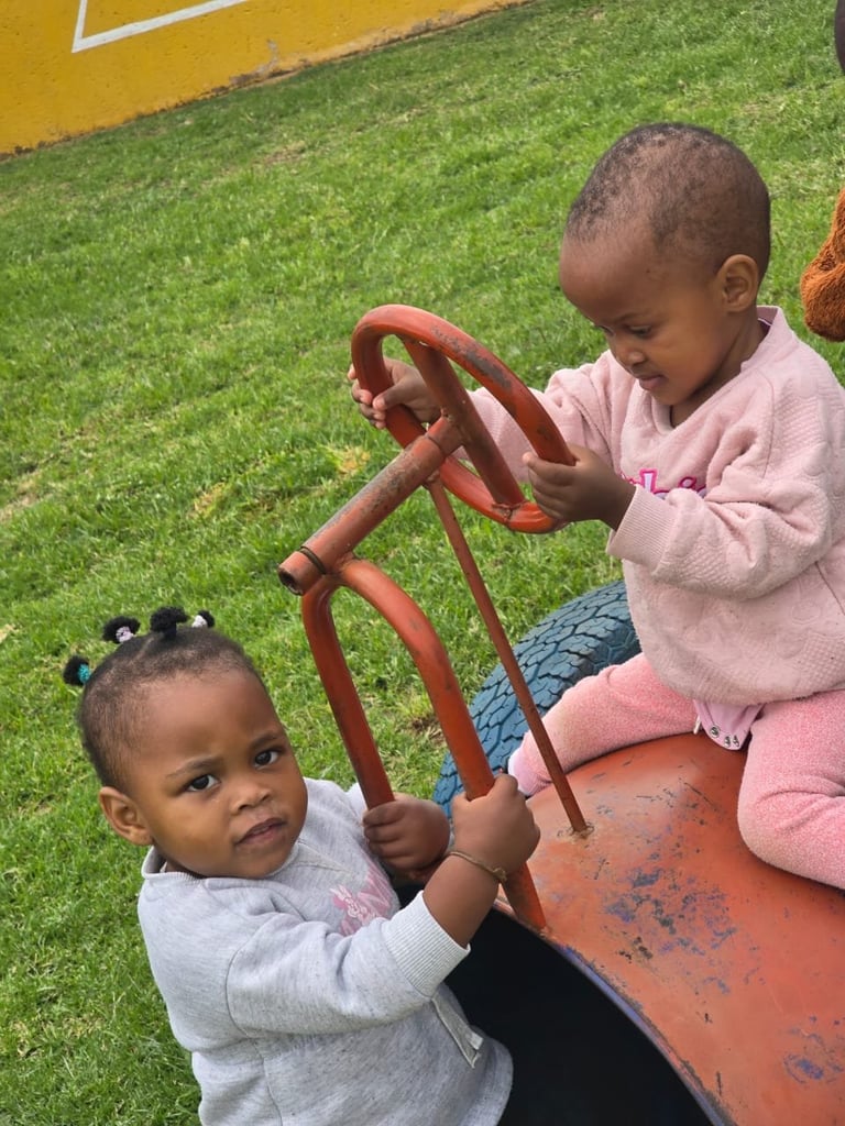 Two toddlers playing on a red playground equipment with green artificial turf and yellow wall in background