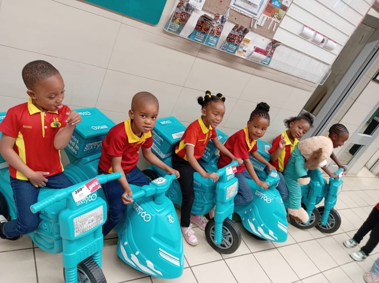 Group of young children in red and yellow uniforms riding blue toy scooters indoors in a classroom or school setting