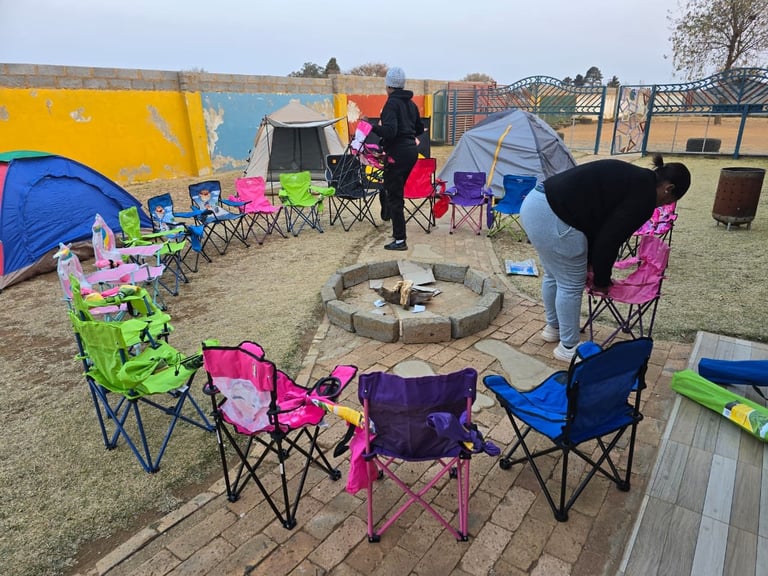 Group of colorful camping chairs arranged in a circle around a fire pit with tents and people at an outdoor campsite