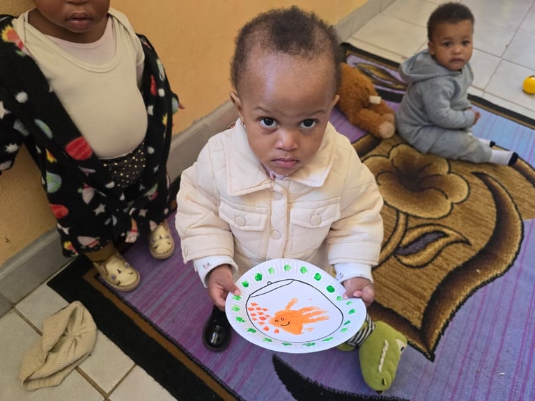 Toddler in white shirt holds a colorful plate with fish design in a daycare or classroom setting with caregiver and other children