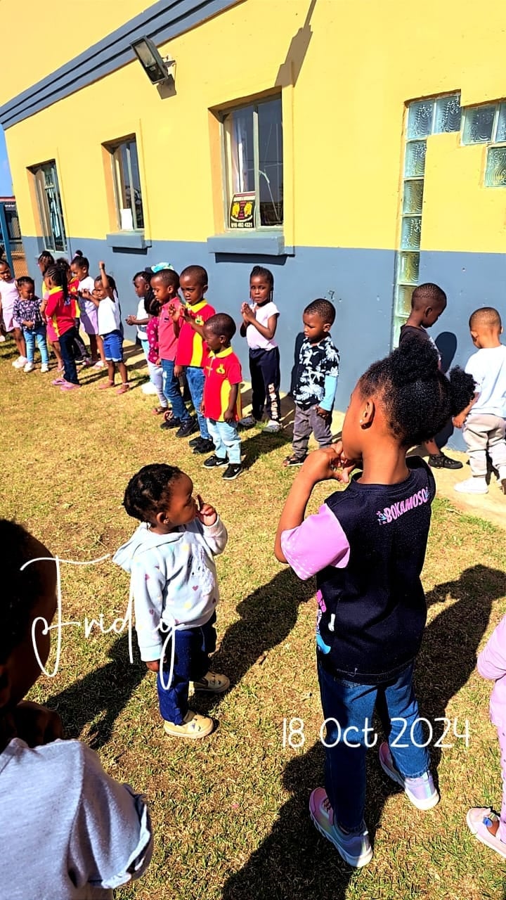 Group of children standing on grass in front of a bright yellow and blue school building outdoors
