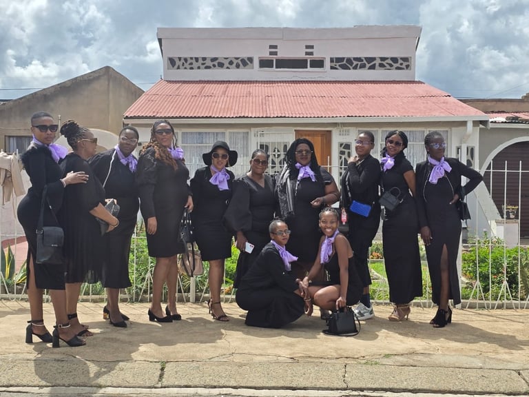 Group of school girls in dark uniforms with purple ribbons standing in front of a building with a pink roof