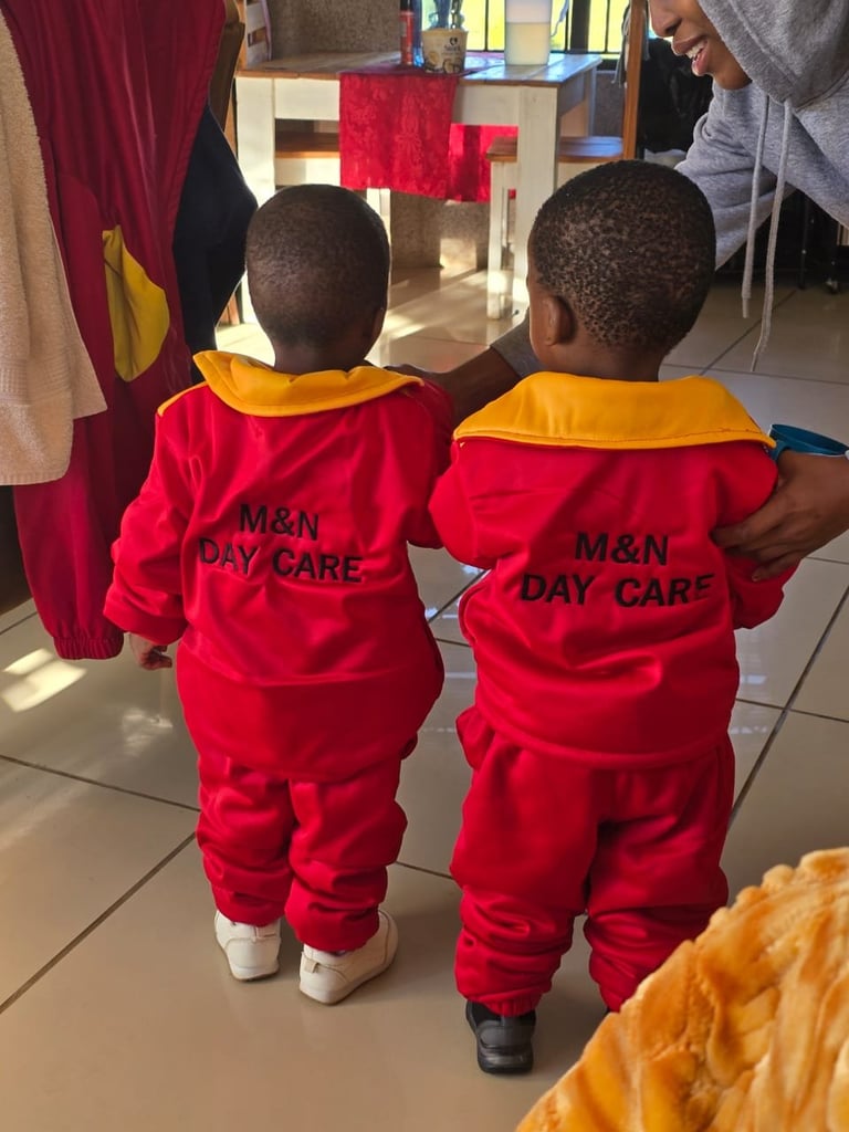 Two young children wearing red and yellow M&N Day Care jackets, viewed from behind in an indoor daycare setting