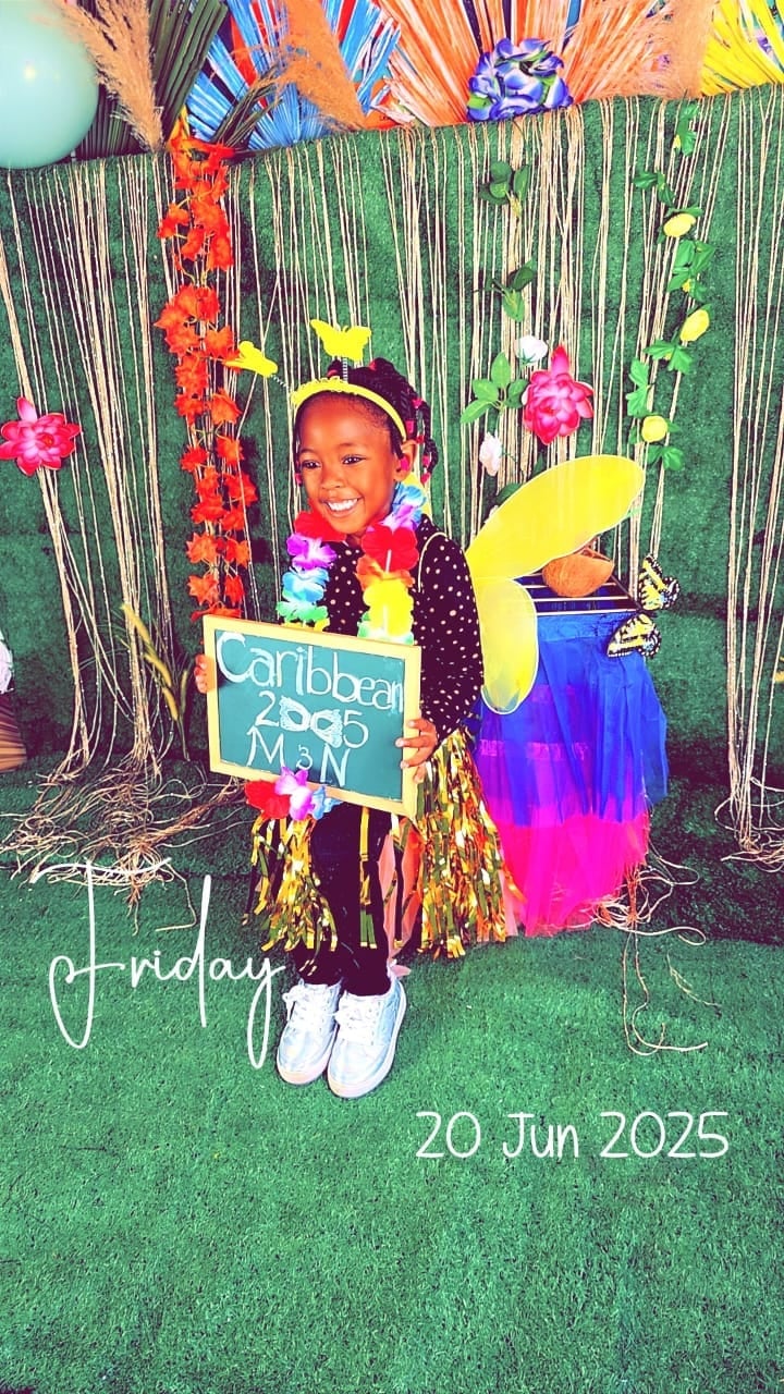Young child in colorful Caribbean-themed outfit posing with tropical decorations and sign reading Caribbeat 2030 Manf, dated June 20, 2025