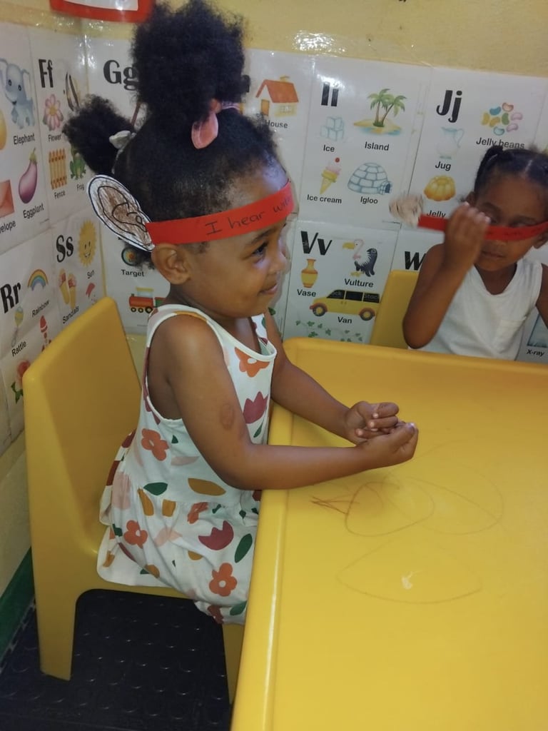 Young child with black hair in red headband sitting at yellow table in classroom with alphabet poster wall
