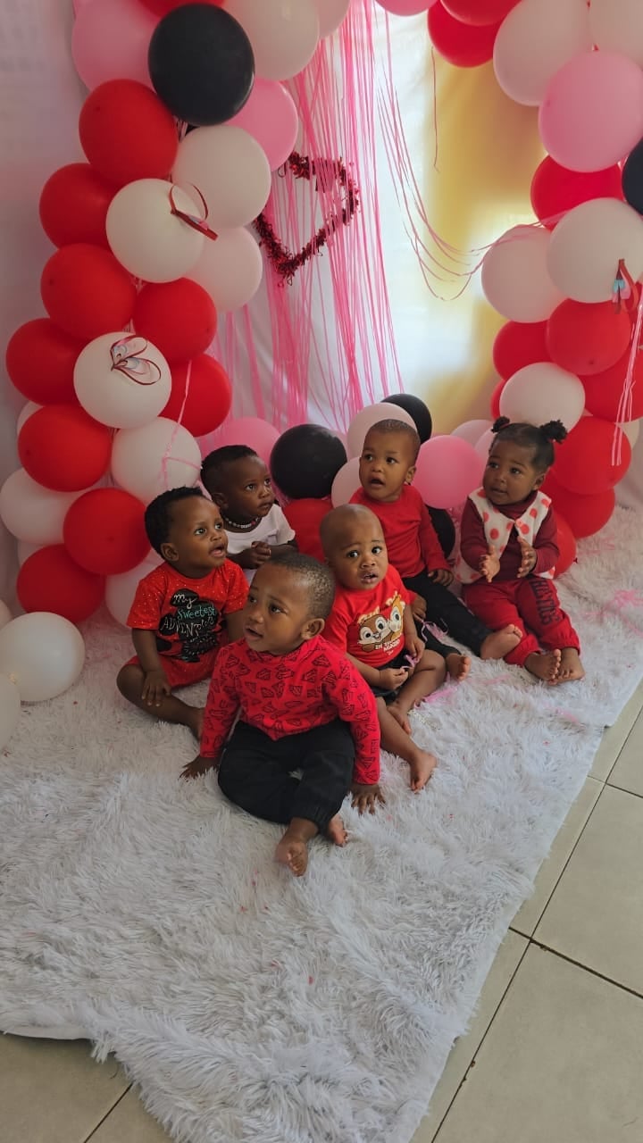 Group of young children sitting on a white fluffy mat surrounded by red, white, pink, and black balloons with heart decorations in background