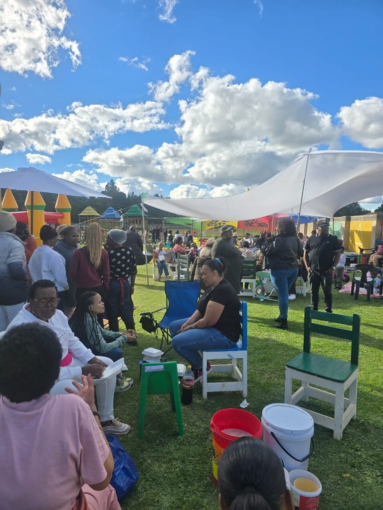 Outdoor community event with colorful tents, people sitting on green benches, and vendor booths under a sunny sky with white clouds