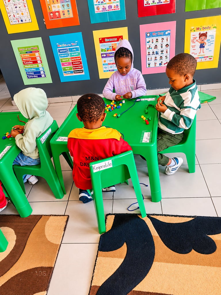 Four young children sitting at a green table playing with colorful beads in a classroom with educational posters on the wall