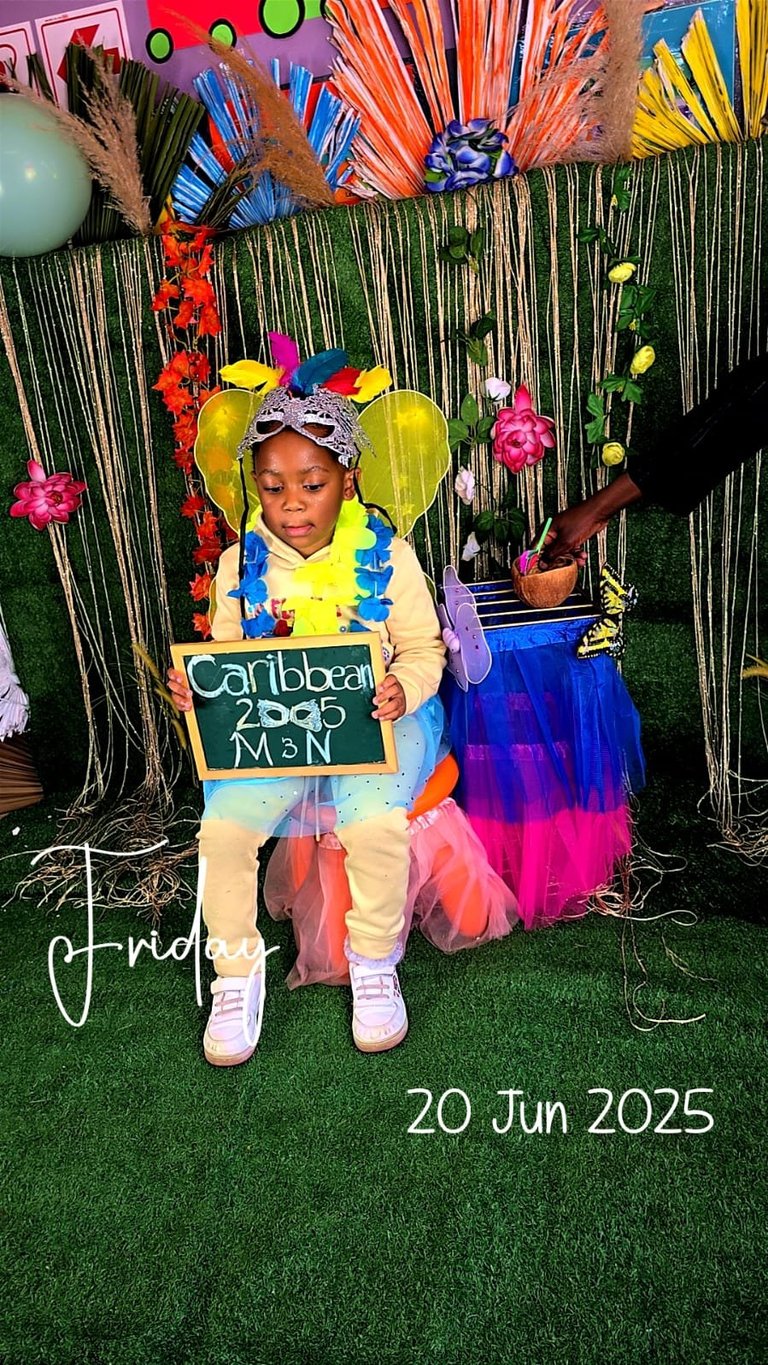 Young child in colorful Caribbean-themed costume holding chalkboard sign at photo backdrop with tropical decorations and date 20 Jun 2025