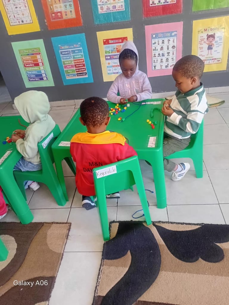 Four young children sit at a green table in a classroom, playing with colorful balls while educational posters hang on the gray wall behind them
