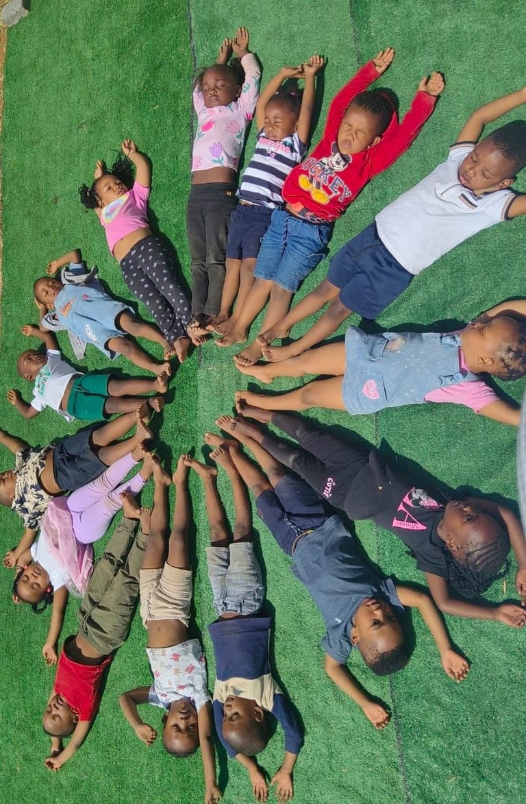 Group of children lying on green grass in a circle formation with hands raised toward center, viewed from above