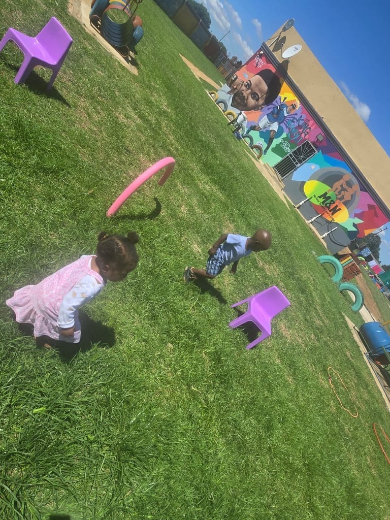 Children playing in a grassy outdoor park area with colorful purple chairs and toys scattered around under a blue sky