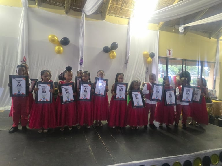 Group of children in red robes holding photos, standing on stage with yellow and black balloons