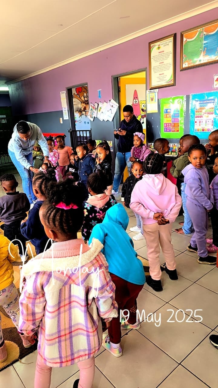 A group of young children gathered in a classroom with pink walls, engaging with teachers during an activity or lesson