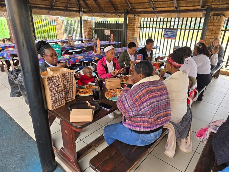 Group of children and adults gathered indoors during a community meal or event, with food and people seated and standing around tables