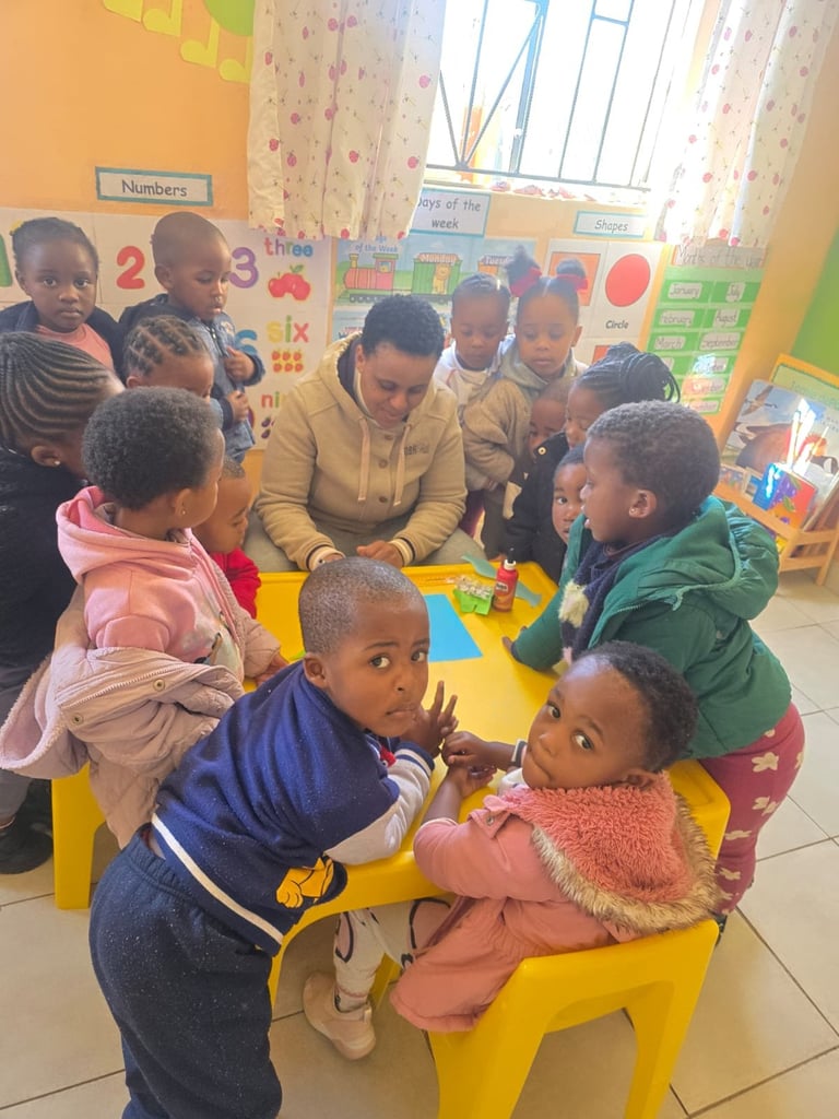 Teacher and young children gathered around a yellow table in a colorful classroom during an interactive learning activity