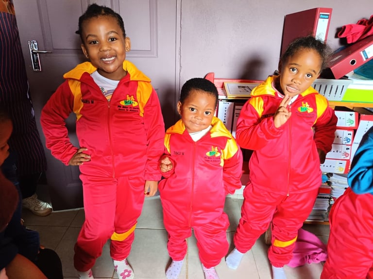 Three young children wearing matching red and yellow jumpsuits smiling in a classroom or daycare setting