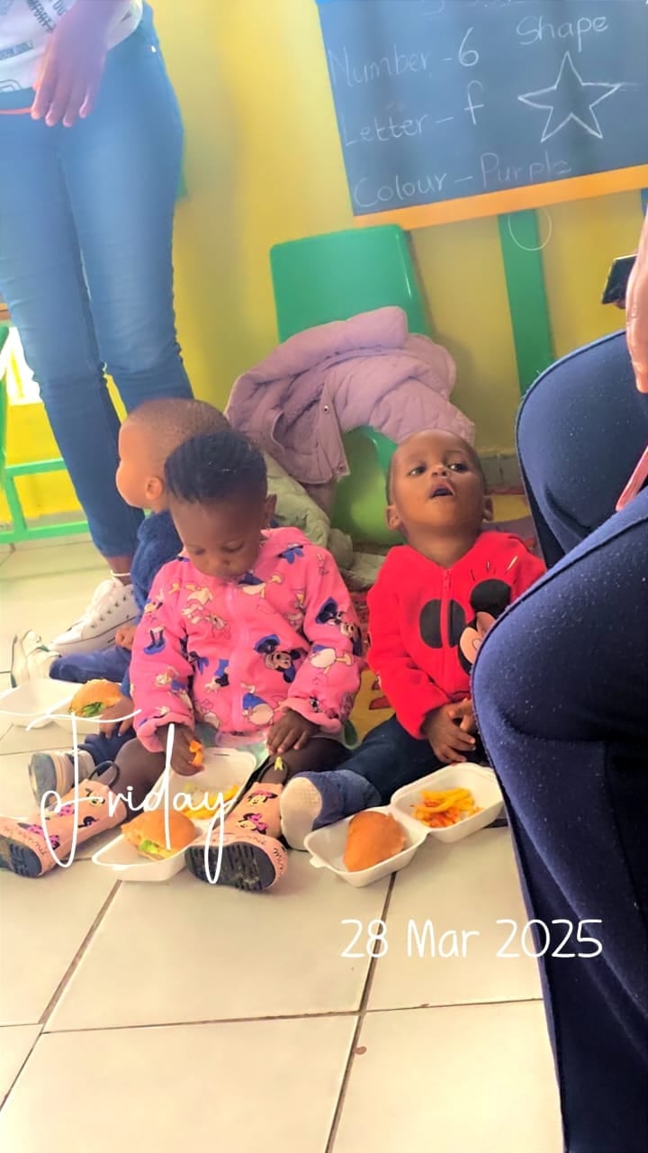 Two toddlers sitting on floor in colorful playroom eating snacks, adults nearby, dated March 28 2025