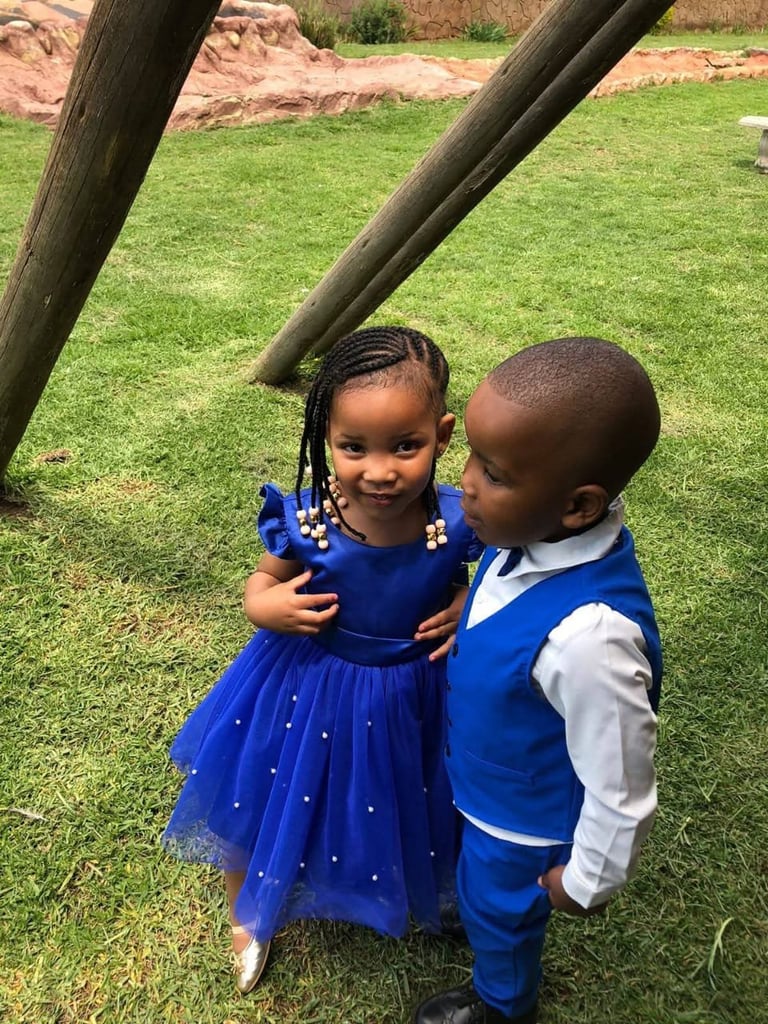 Two young children in formal blue outfits posing together on green grass under tree branches