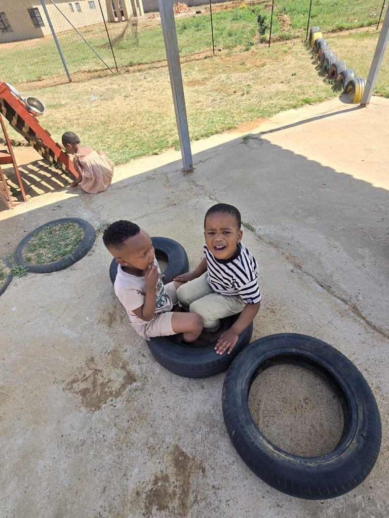 Two young children playing with black tires in a concrete outdoor play area with metal fence and grass field visible in background