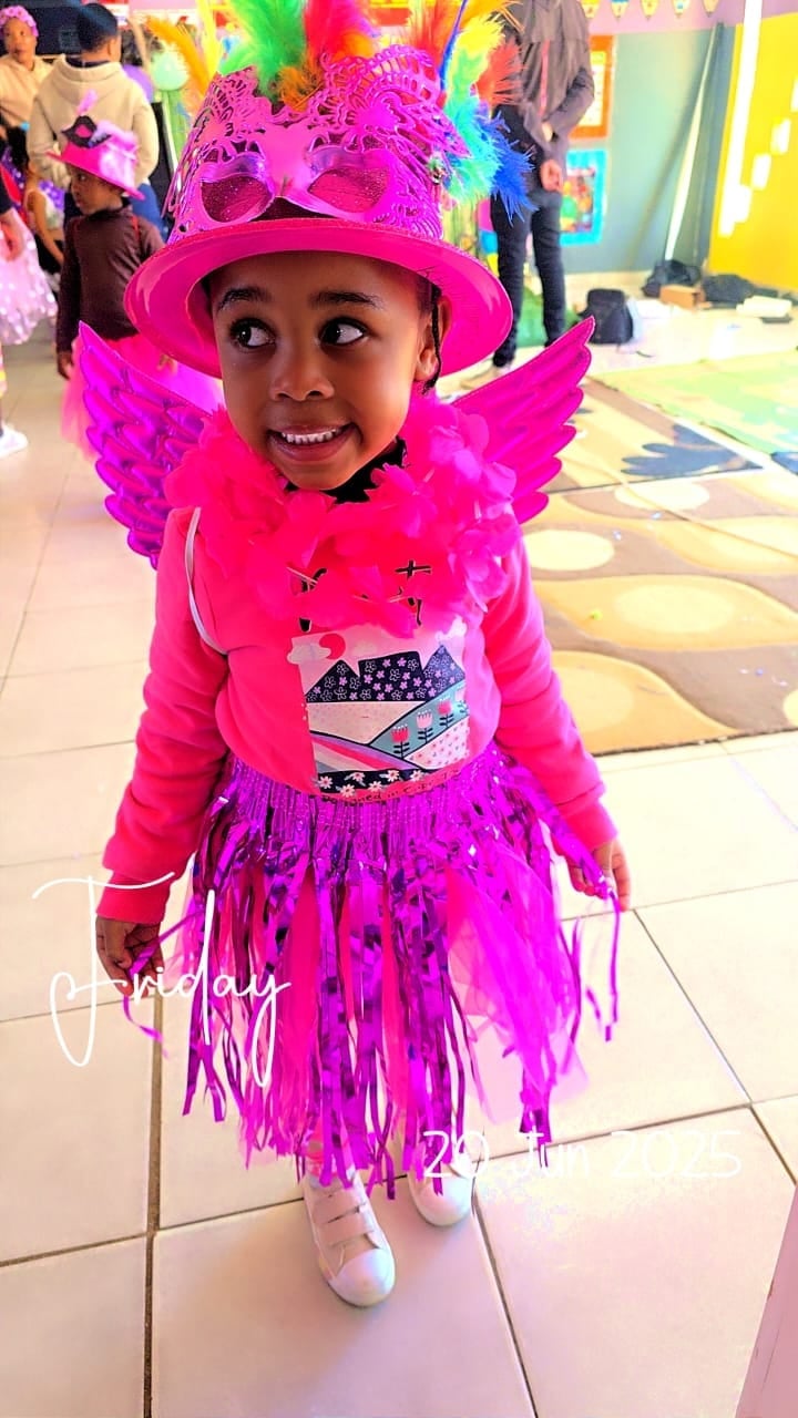 Child wearing bright pink and purple costume with feathered wings, sequined hat, and fringed skirt at colorful indoor party event