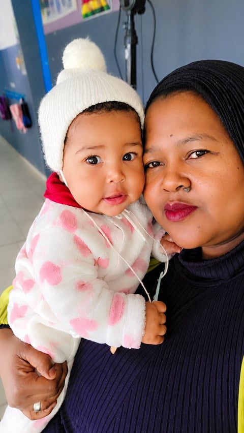 Adult woman holding a baby wearing a white beanie and pink polka dot outfit, both smiling at camera indoors
