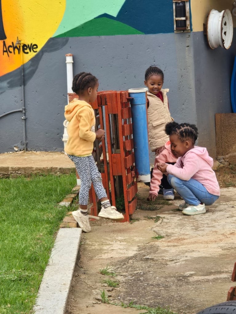Three young children playing with a red wooden structure at an outdoor playground with colorful mural backdrop