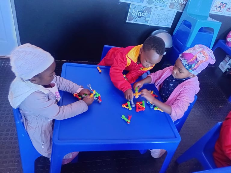 Three young children playing with colorful building blocks at a blue table in a classroom setting