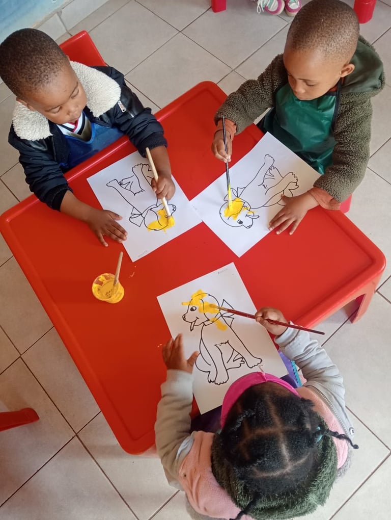 Three young children painting yellow designs on coloring pages at a red table during an art activity