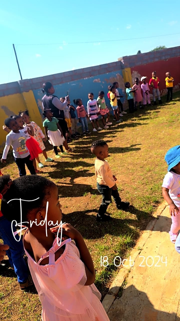 Large group of children in colorful clothing lined up outdoors next to a blue and red building on a sunny day