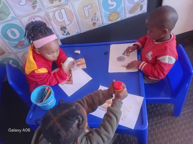 Three young children sitting at a blue table engaging in an art or craft activity with papers and materials