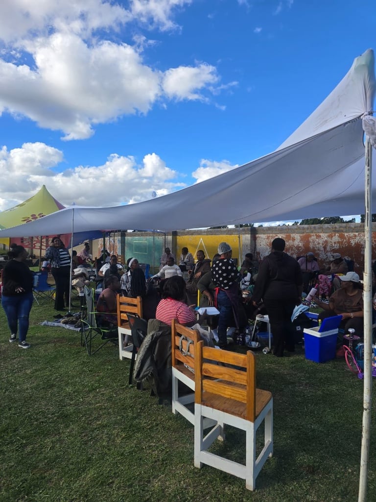 Outdoor event under white tent canopy with colorful chairs and multiple attendees gathered in grassy field on sunny day