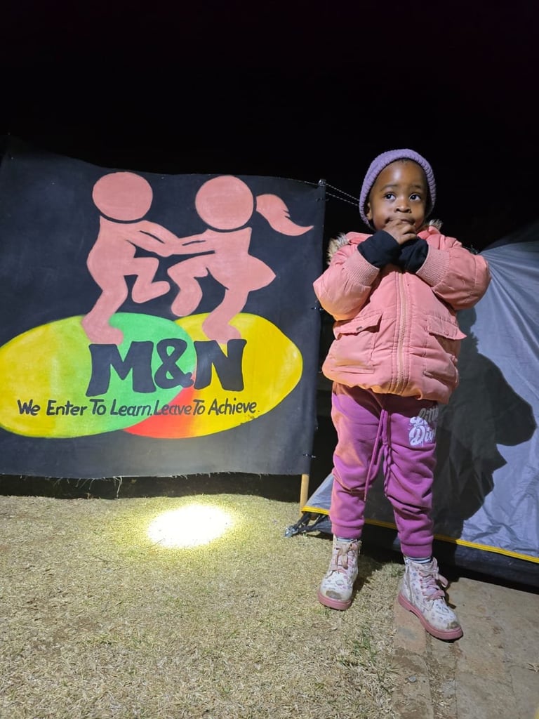 Young child in pink jacket and purple pants standing in front of M&N educational organization banner at night