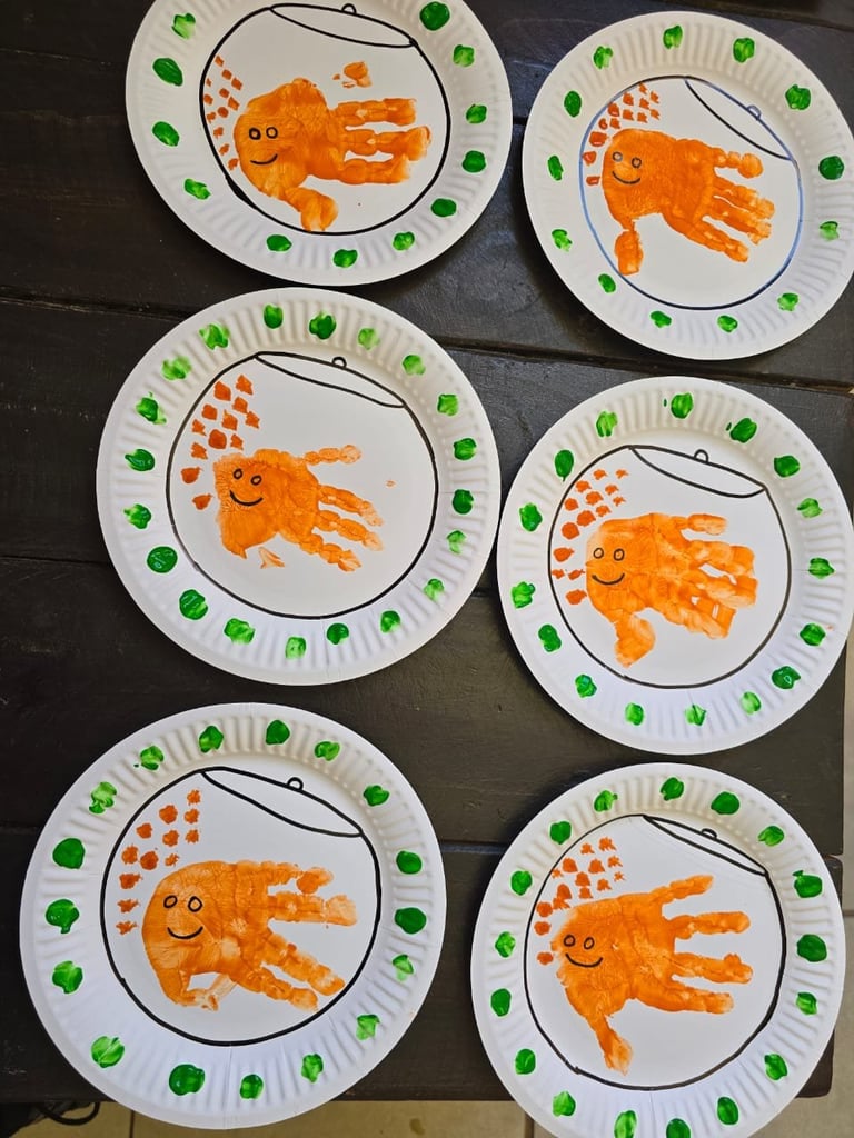 Six paper plates with handprint caterpillar crafts made from orange and green fingerprints arranged on a dark surface