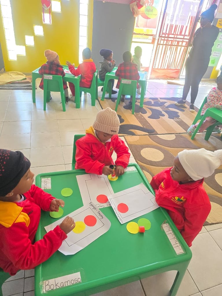 Young children in red jackets doing color-matching activities at green tables in a bright classroom with yellow walls