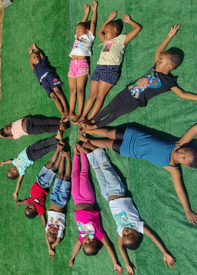 Group of diverse children lying on green grass in a circle with hands joined at center, viewed from above