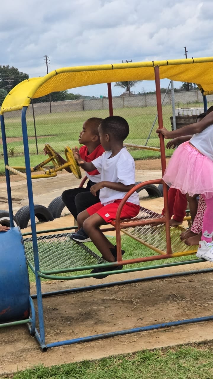 Children riding a colorful manual merry-go-round on a dirt playground with green grass and fenced fields in the background