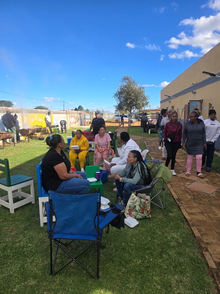 Community outdoor event with people gathered on grass, sitting in camp chairs and standing near a brown building on a sunny day